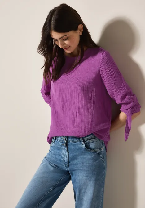 Frau mit lila Bluse und blauer Jeans lehnt an einer Wand.